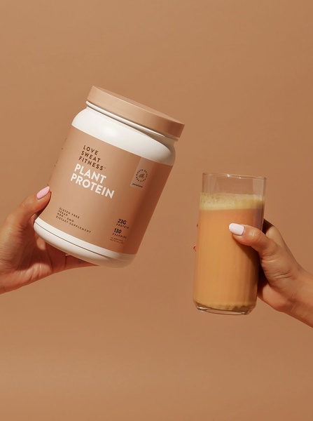 A high-contrast, clean-aesthetic product shot showing a hand holding a container of plant protein next to a glass of the prepared drink.